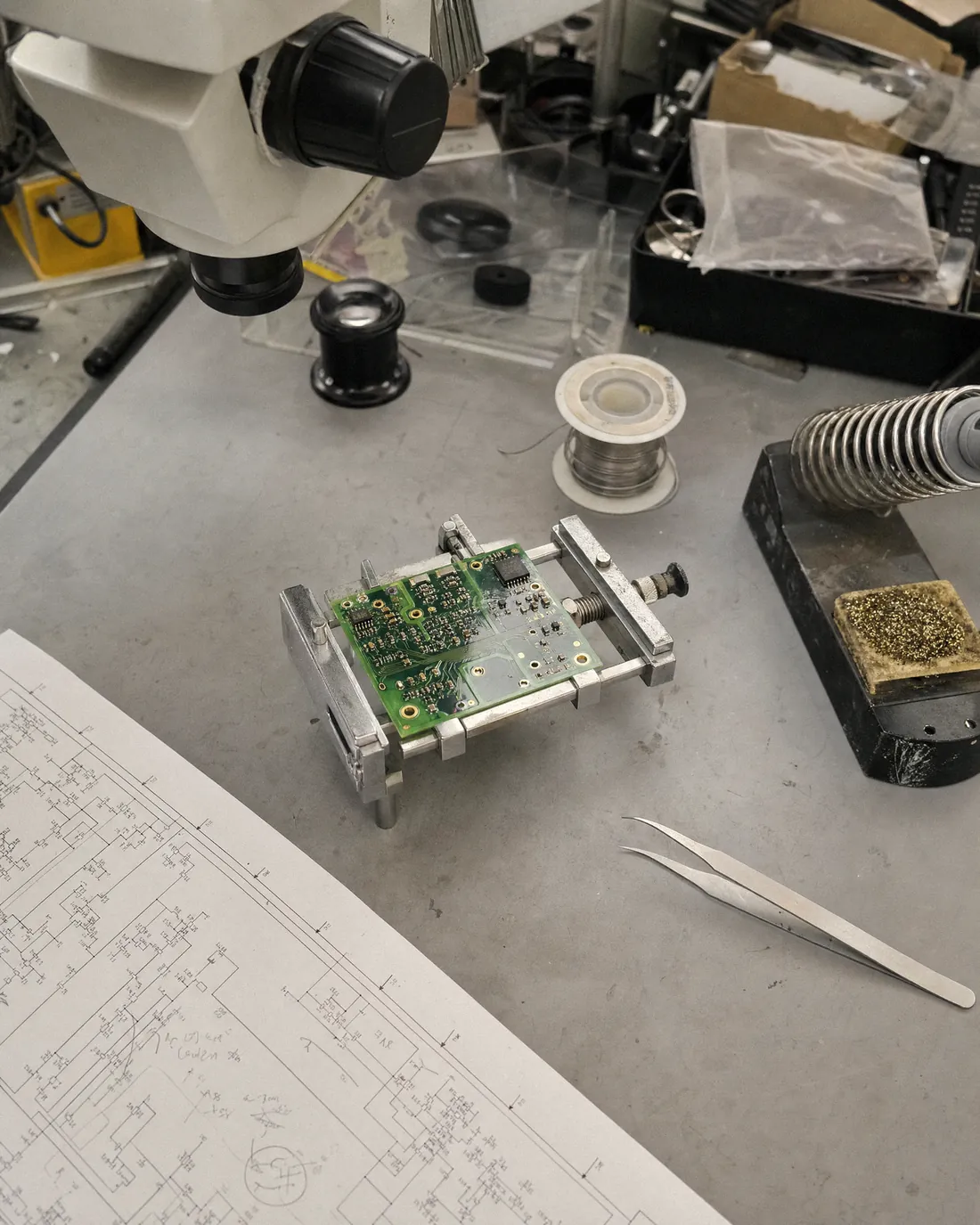 A small green circuit board clamped in a metal holder on a grey bench mat, with tweezers, a soldering iron stand, solder and a printed schematic marked up in pencil.