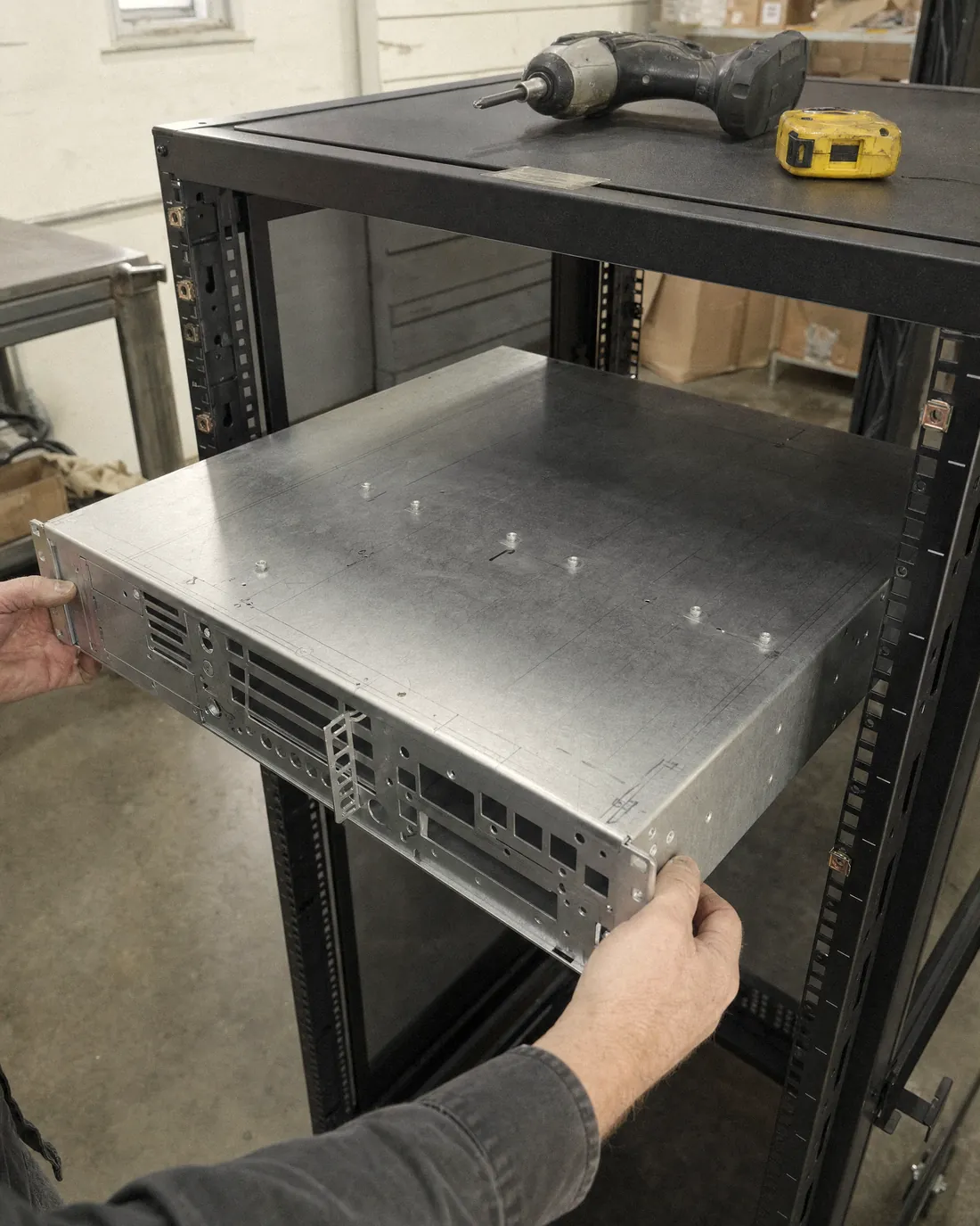 Two hands sliding an unpainted sheet-metal 2U server chassis into a black equipment rack. Pencil layout lines are still on the metal. A tape measure and a cordless driver sit on top of the rack.